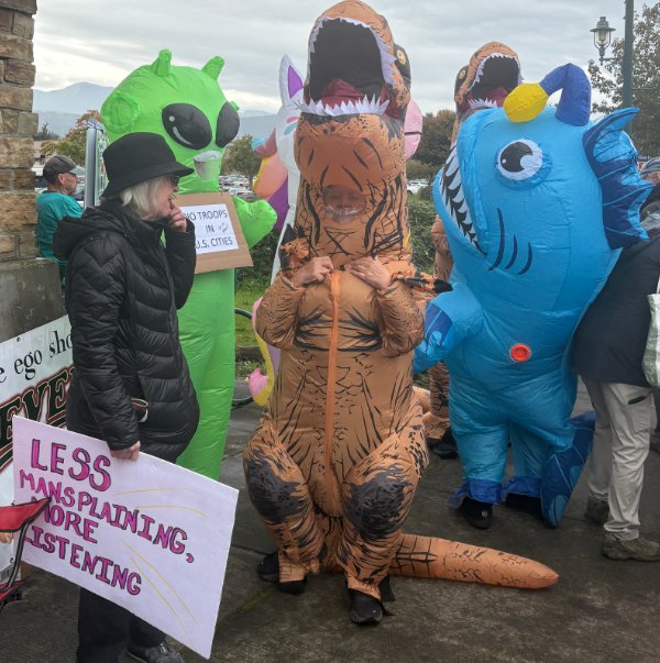 Get together with blow up dinosour, shark, and alien with human carrying sign Less Mansplaining More Listening